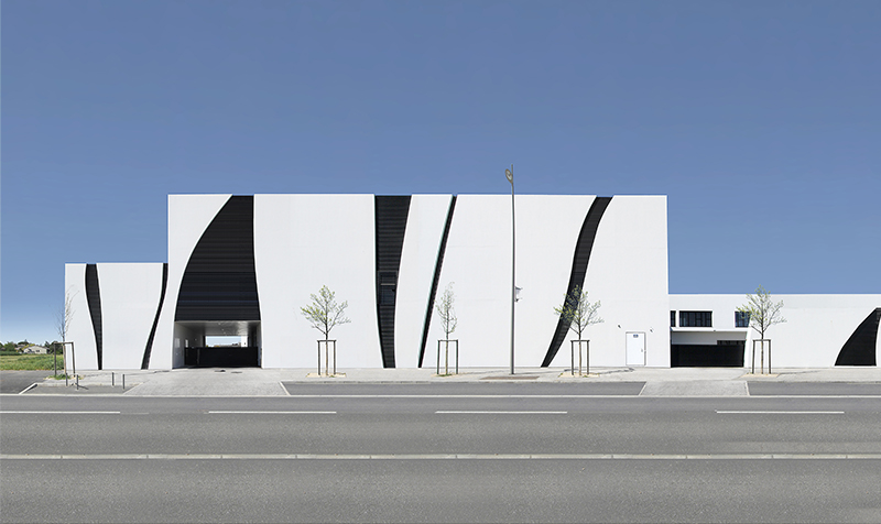 Photographie de la 1ere partie d'un panoramique de la facade principale depuis la rue, bâtiment béton blanc avec des arabesques noires tout hauteur comme des tiges grimpant vers le ciel