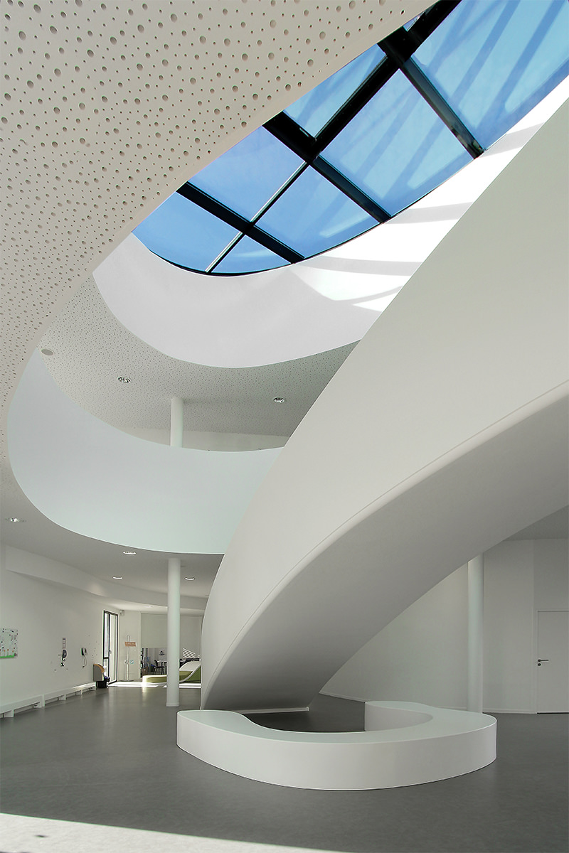 Photographie vertical du hall d'accueil immaculé, un puits de lumière éclaire les deux étages et l'escalier situé au centre