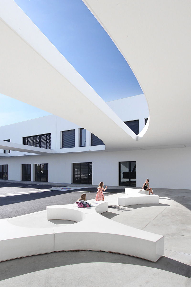 Photographie vertical de la cour d'école, en dessous des deux auvents courbe, on voit la facade noir et blanc, des enfants sont sur des bancs aux formes abstraites