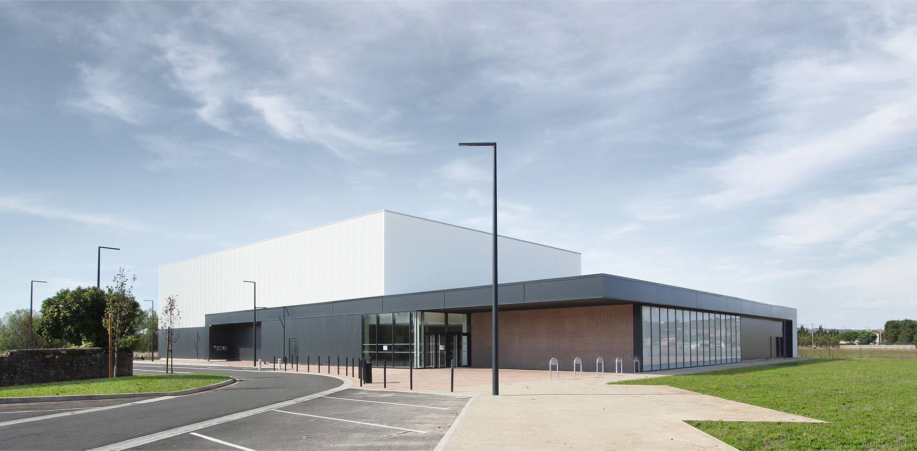 Photographie de l'ensemble du gymnase depuis le trottoir ménant à l'entrée, auvent qui fait le tour du bâtiment gris anthracite et soubassement brique, volume bardage blanc vertical pour le terrain couvert du gymnase