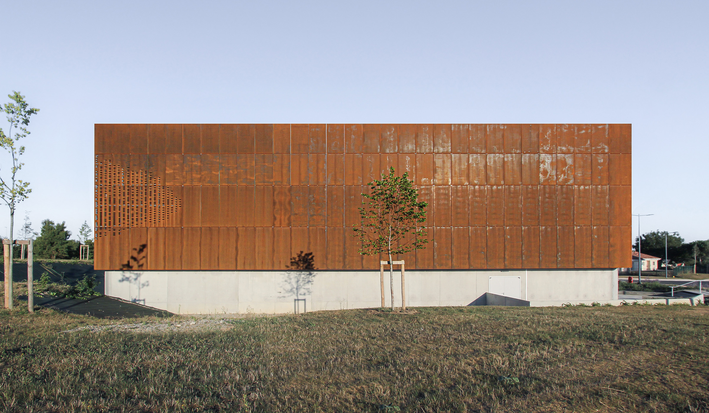 Photographie de la facade en corten du gymnase de l’Isle-Jourdain, soubassement en béton, entouré d'espace vert