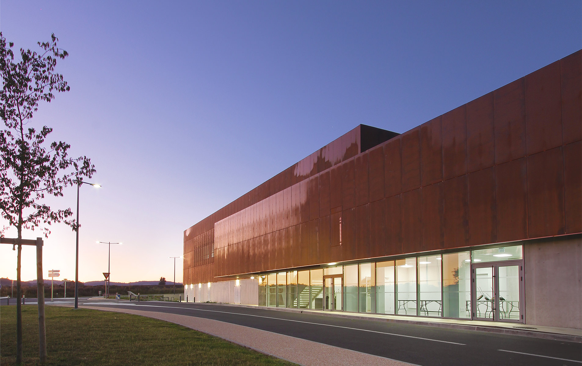 Photographie de l'ensemble du gymnase depuis l'espace à côté de l'entrée, soubassement en béton brut et corten oxydé