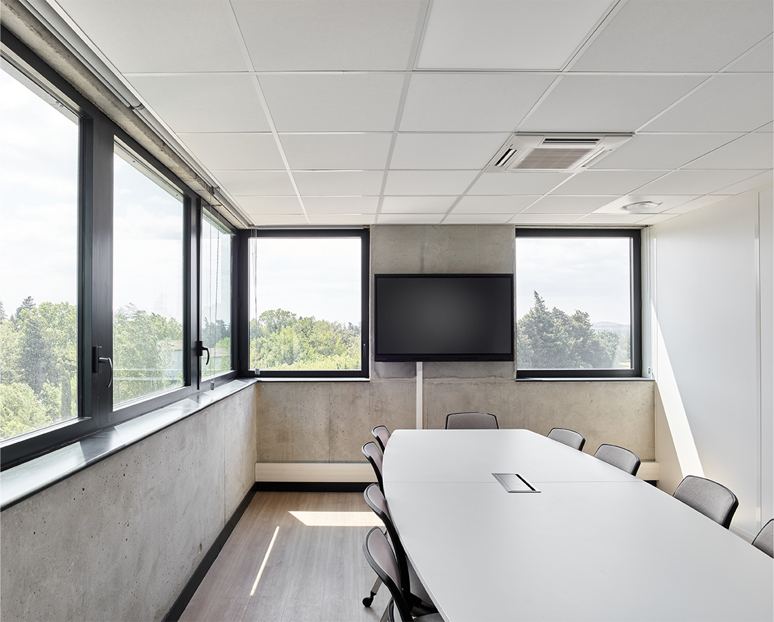 Photographie d'une salle de réunion sur un angle du bâtiment, fenêtres bandeaux gris anthracite et soubassement béton brut, plancher bois et murs blanc