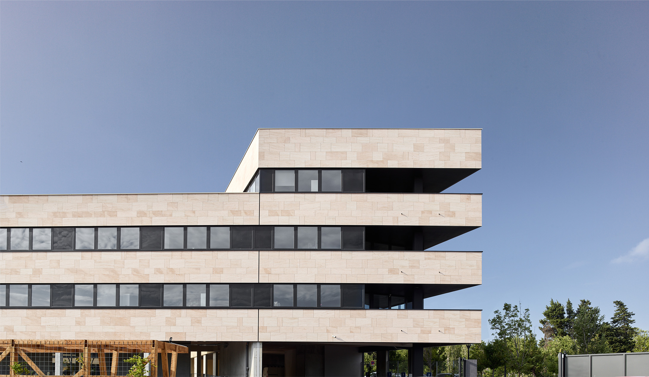 Photographie de la façade arrière, alternance en étage des murs en pierre beige et des fenêtres bandeaux gris anthracites, fond arboré et ciel bleu clair