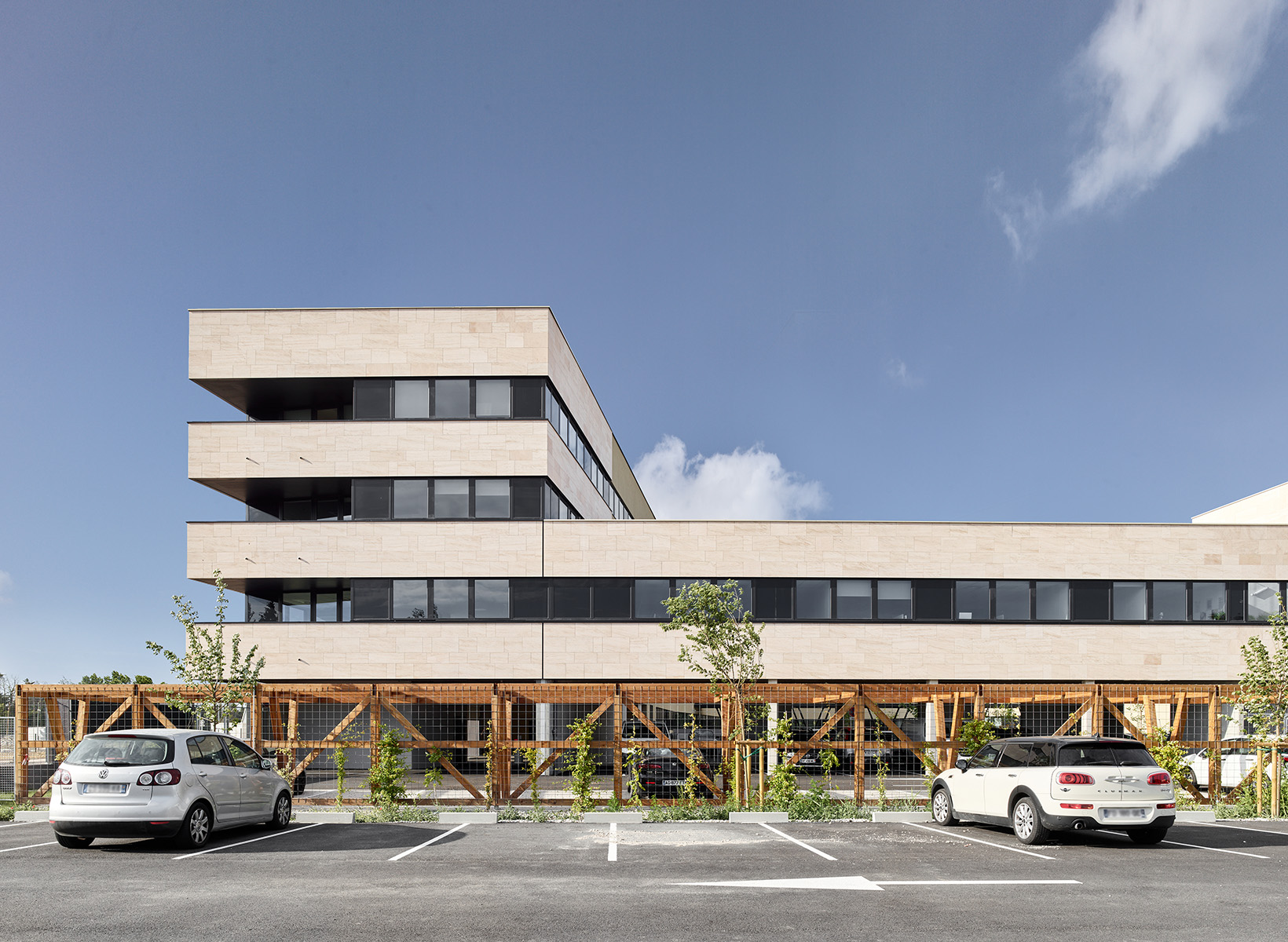 Photographie face aux place de parking et au bâtiment, entre, une pergola commence à voir sa végétation pousser, bâtiment fenêtre bandeau gris et pierre beige