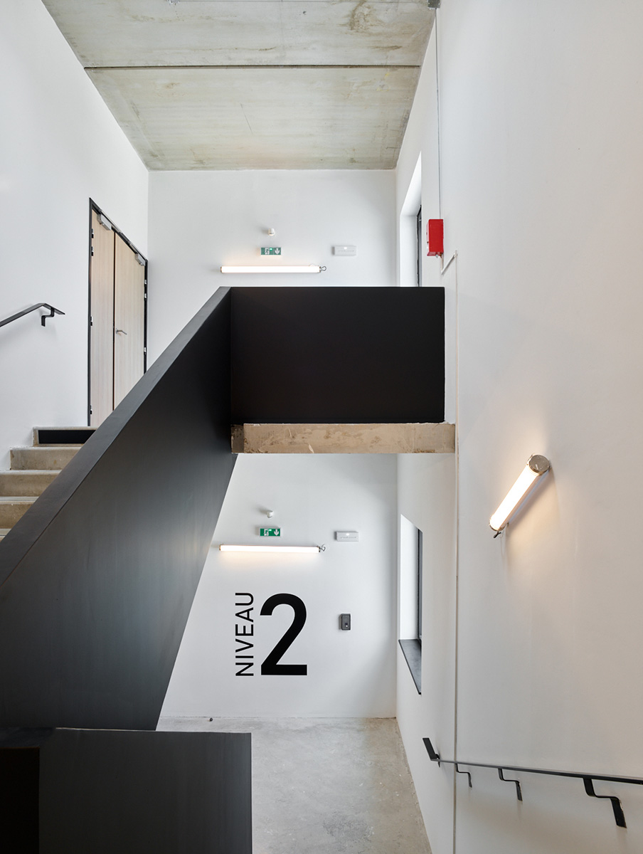 Photographie d'une cage d'escalier avec rambarde mur noire, béton brut sol et plafond et murs blanc , menuiseries gris anthracite