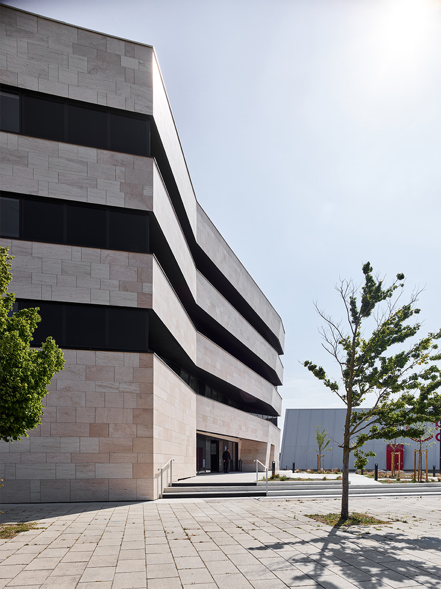 Photographie vue de côté de l'entrée du bâtiment vêtu de pierre beige, fenêtre bandeau sombre qui continu sur le côté, décallage de toute la hauteur de l'entrée de la façade en angle obtu ouvert vers le parvis