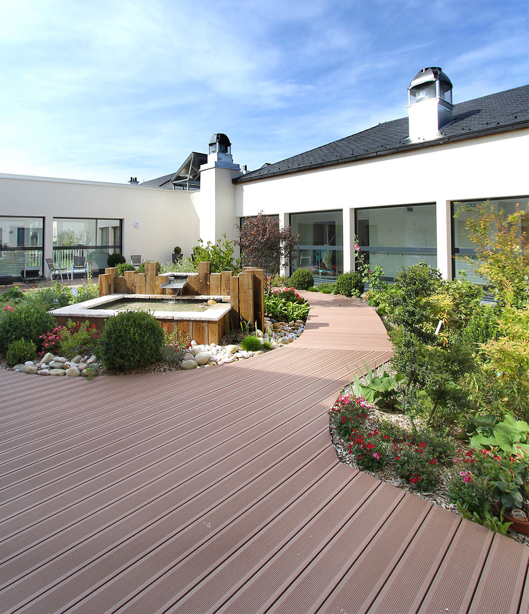 Photographie du patio intérieur avec plusieurs aménagement fleurals, fontaine d'eau et cheminement bois