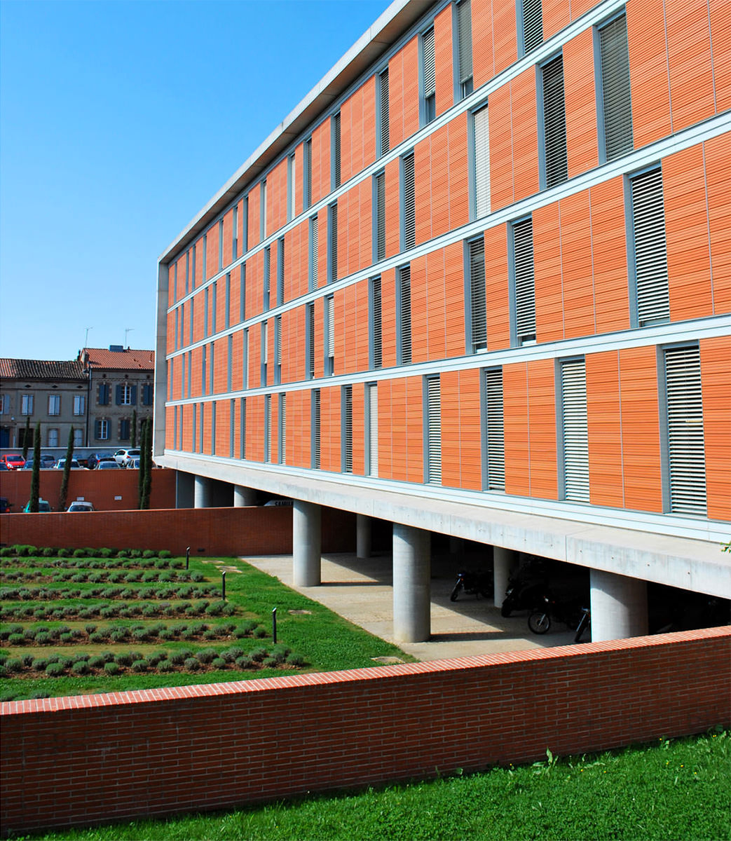 Photographie de biais de la façade sur pilotis et alternance de vitrage et panneaux horizontaux couleur brique sur quatres étages