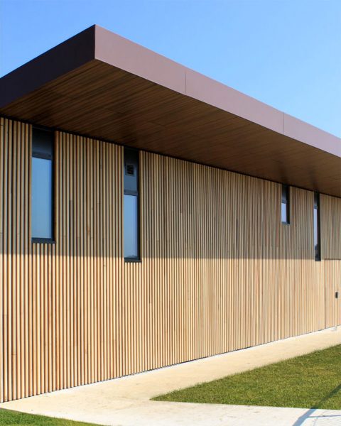 Photographie verticale du bâtiment avec des lames en bois et ouvertures vitrées de hauteurs variables, auvent couleur brune