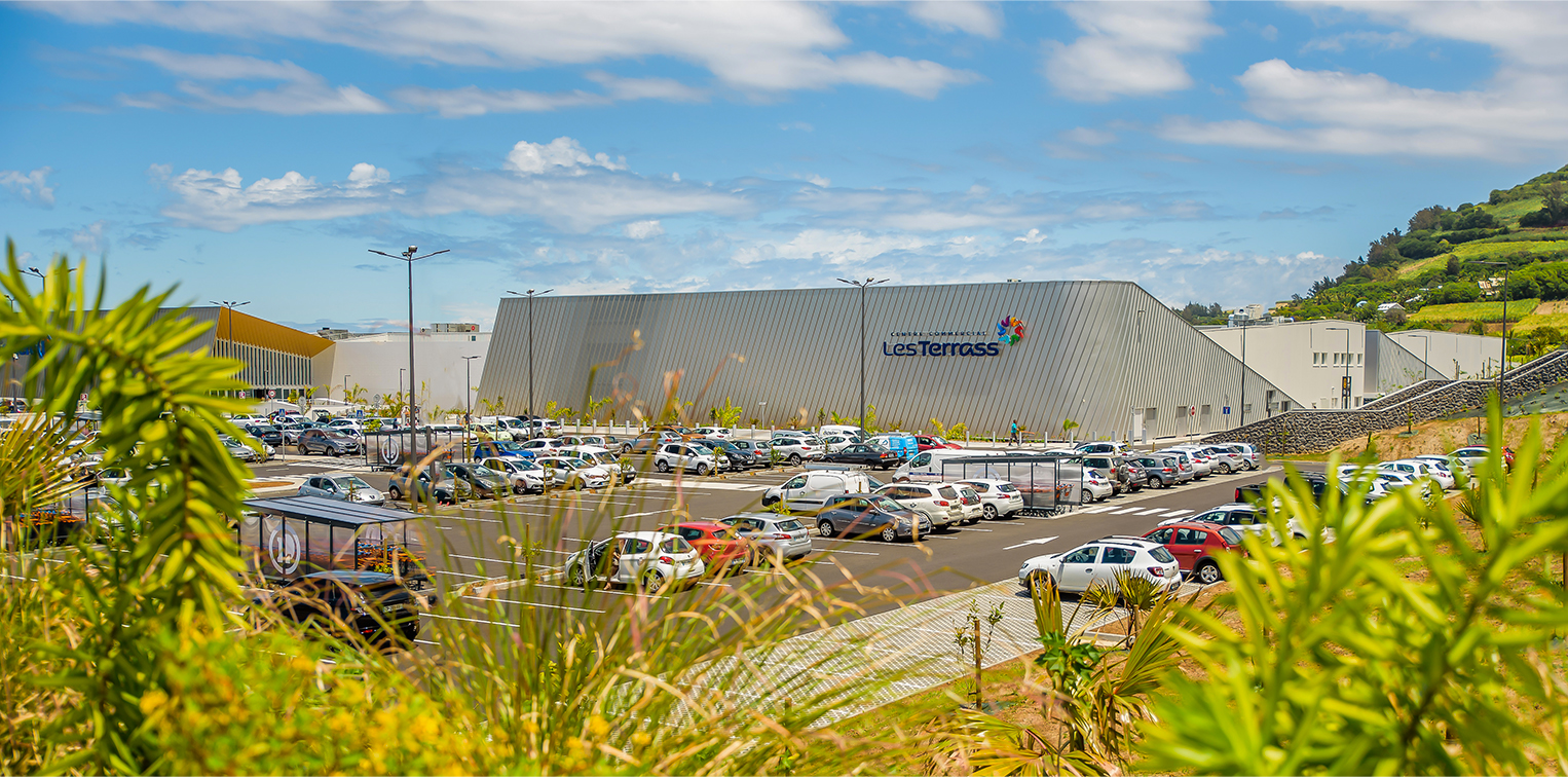 Photographie de jour depuis les hauteurs surplombant le parking, vue sur une partie du centre commercial bardage gris de biais