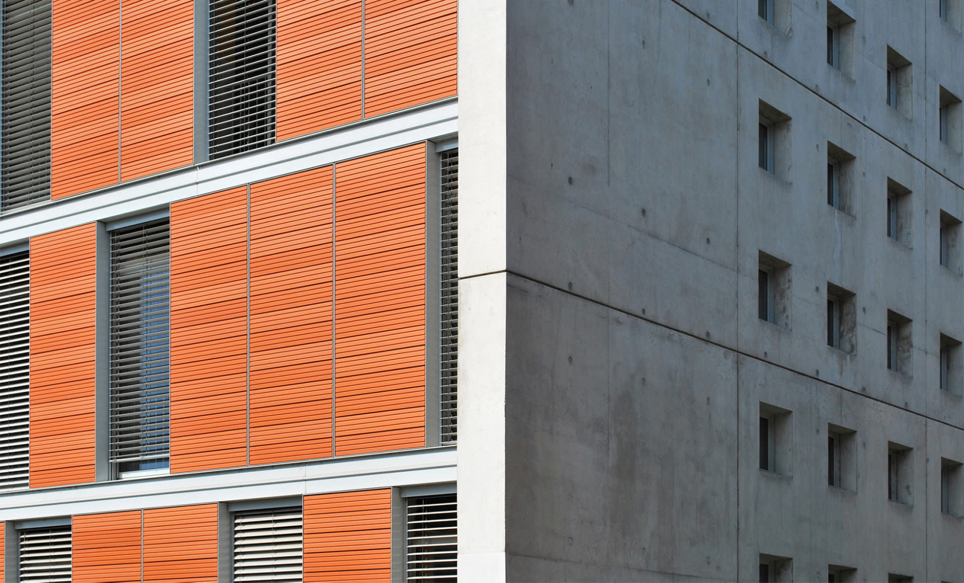 Photographie d'une arrête du bâtiment, d'un côté la partie béton avec de petites ouvertures fenêtres et de l'autres des grand panneaux couleur brique avec des ouvertures fenêtres avec store brise soleil