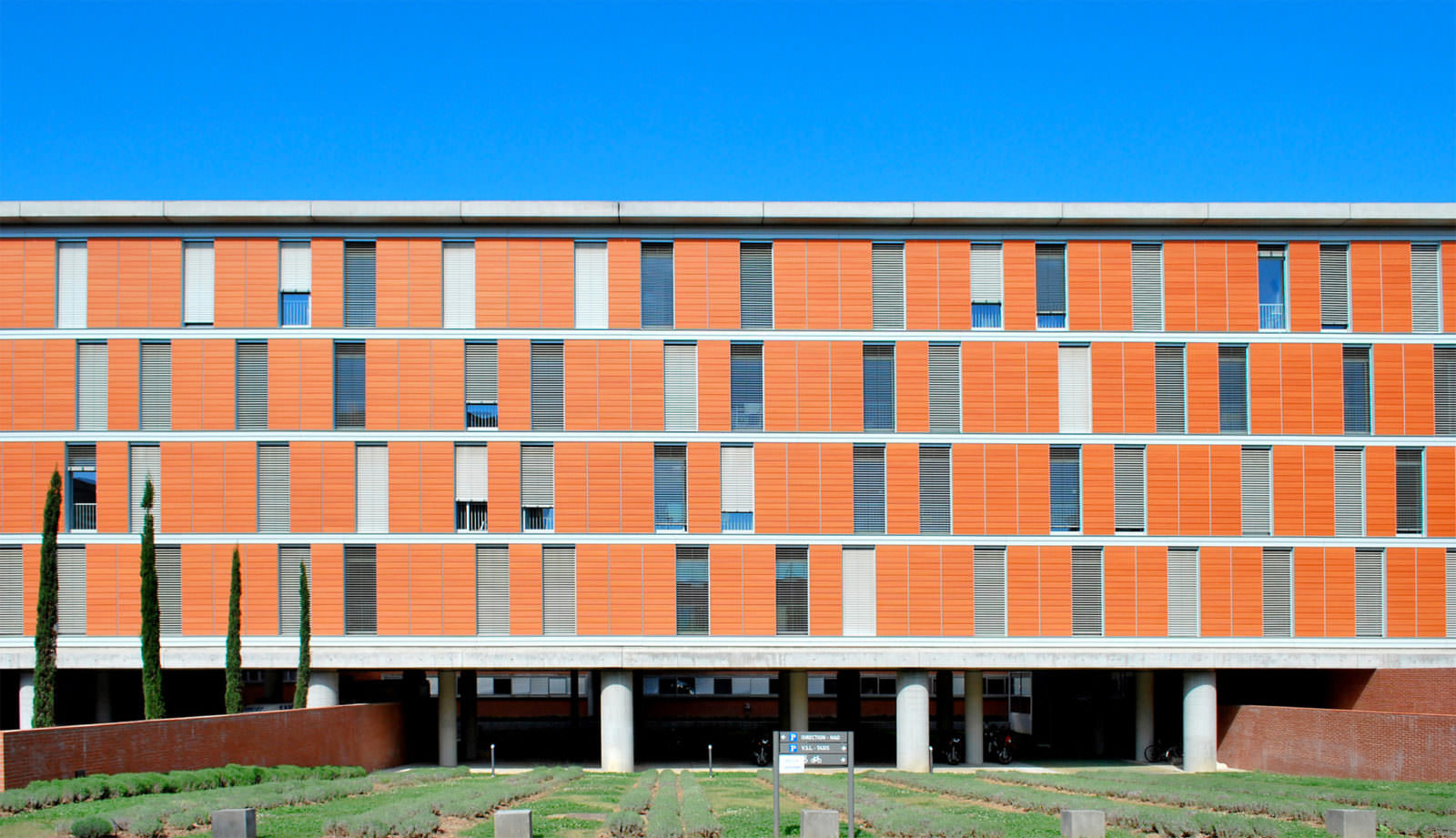 Photographie de la façade principale depuis la rue, espace vert en rangé de massifs avant le bâtiment béton sur pilotis, quatres niveaux en alternance de vitrage et de volet brique