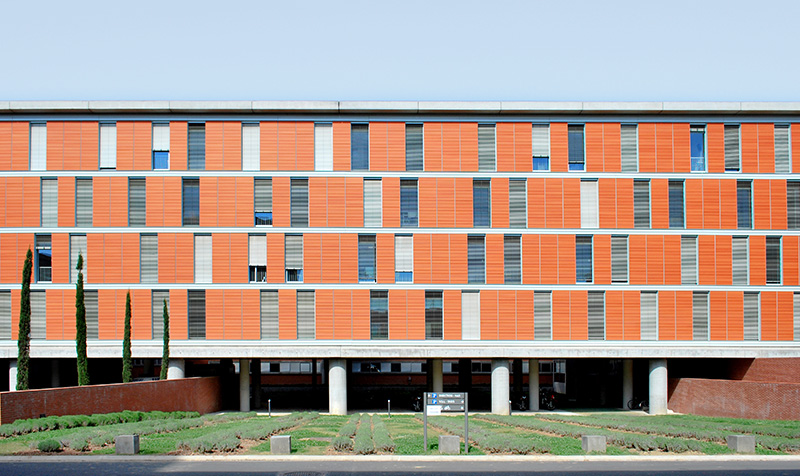 Photographie de la façade principale depuis la rue, espace vert en rangé de massifs avant le bâtiment béton sur pilotis, quatres niveaux en alternance de vitrage et de volet brique