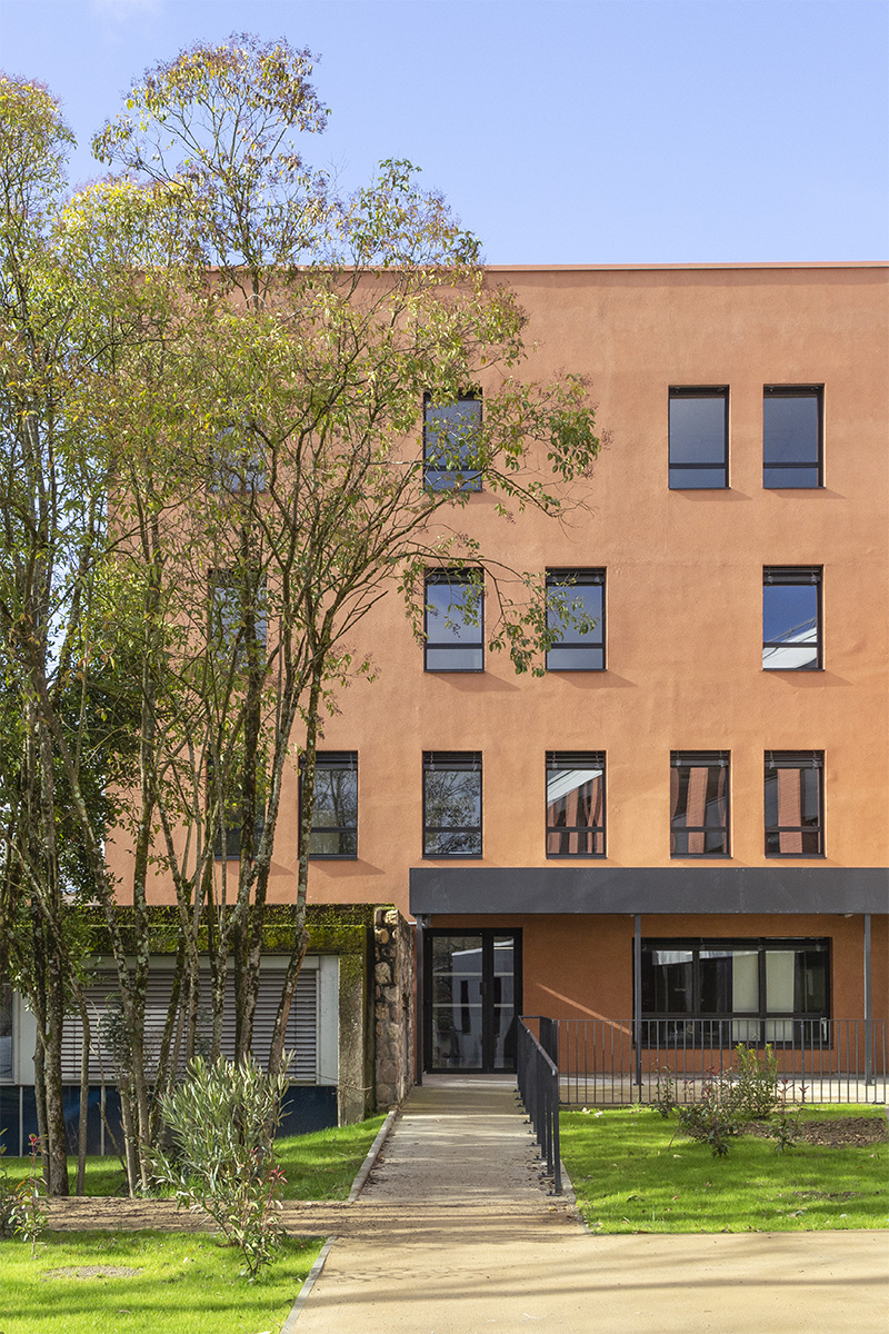 Photographie de l'arrière-cour du bâtiment teinte brique sur 4 niveaux, auvent gris anthracite au 1er niveau, quelques arbres devant le bâtiment