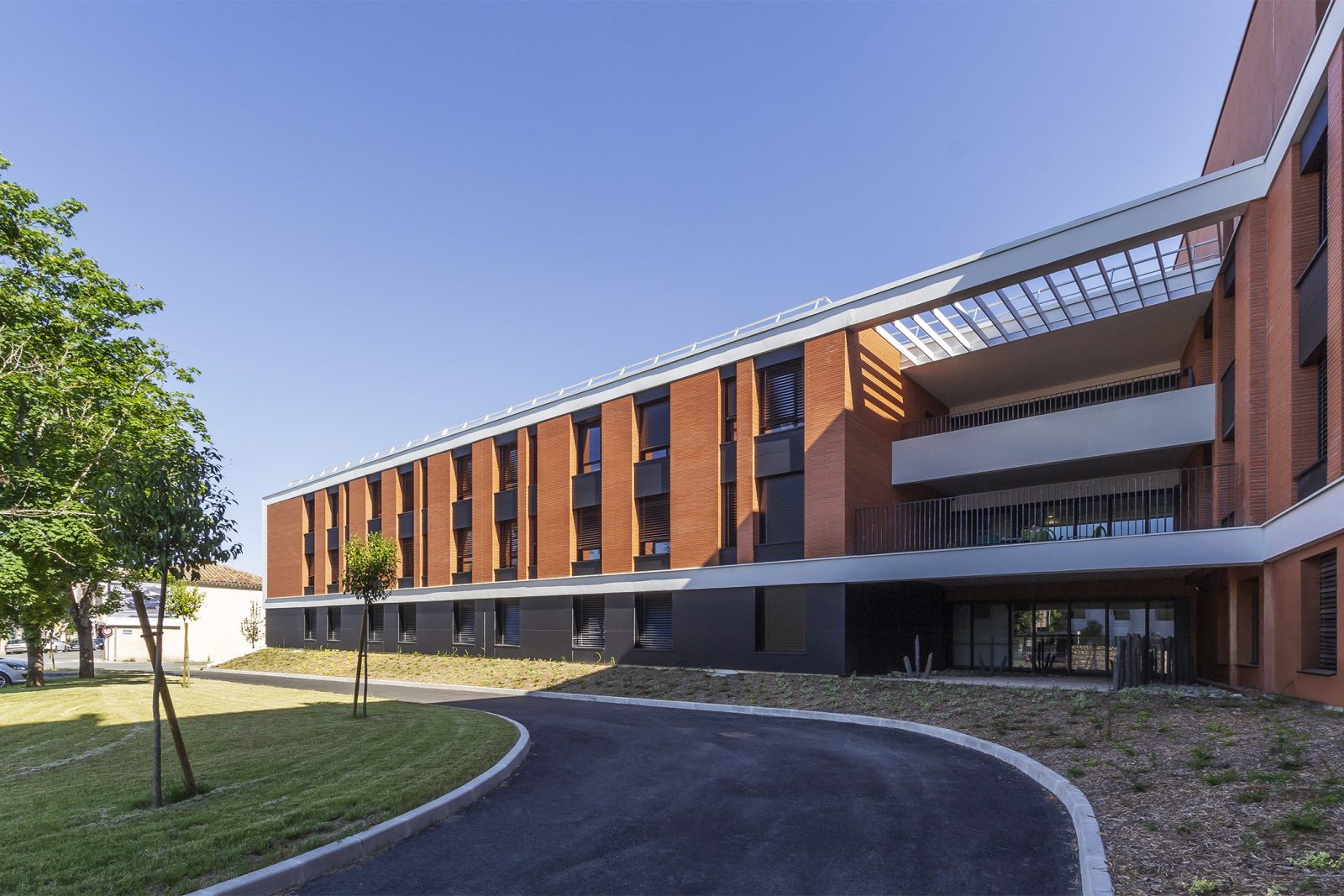 Photographie du centre hospitalier de Gaillac depuis l'accès vehicule dédié à l'hôpital, vue sur le bâtiment en brique tout en longueur, encadrement blanc du niveau 2 et 3 en façade en soubassement gris anthracite