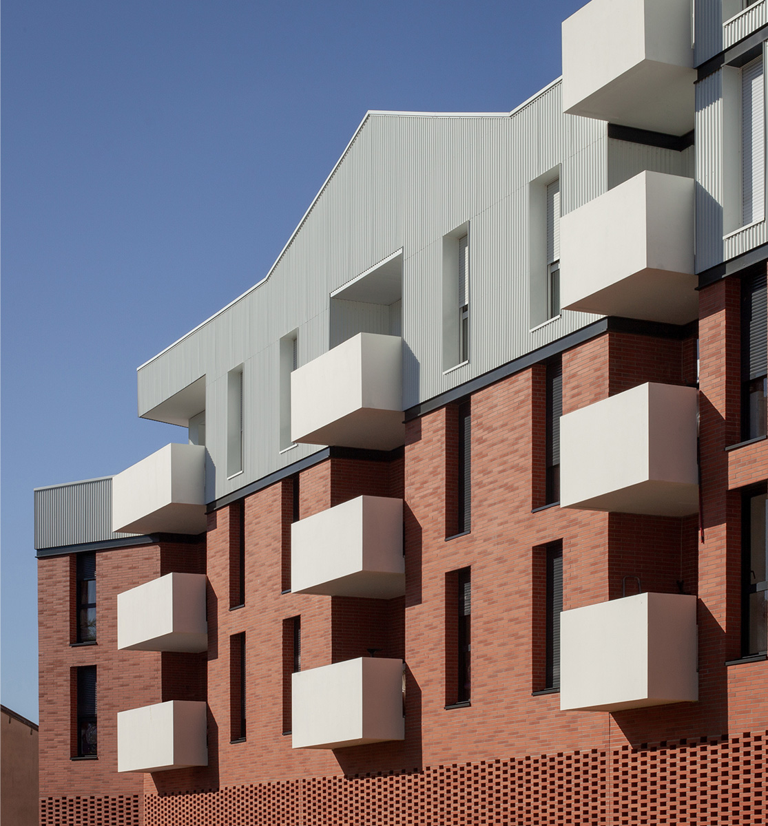 Photographie cadré sur une partie de la façade avec 3 rangées de balcons blanc alignées hozitontalement, briques panachées avec moucharabieh en partie basse (jusqu'a R+2) puis bardage vertical métallique blanc en partie haute