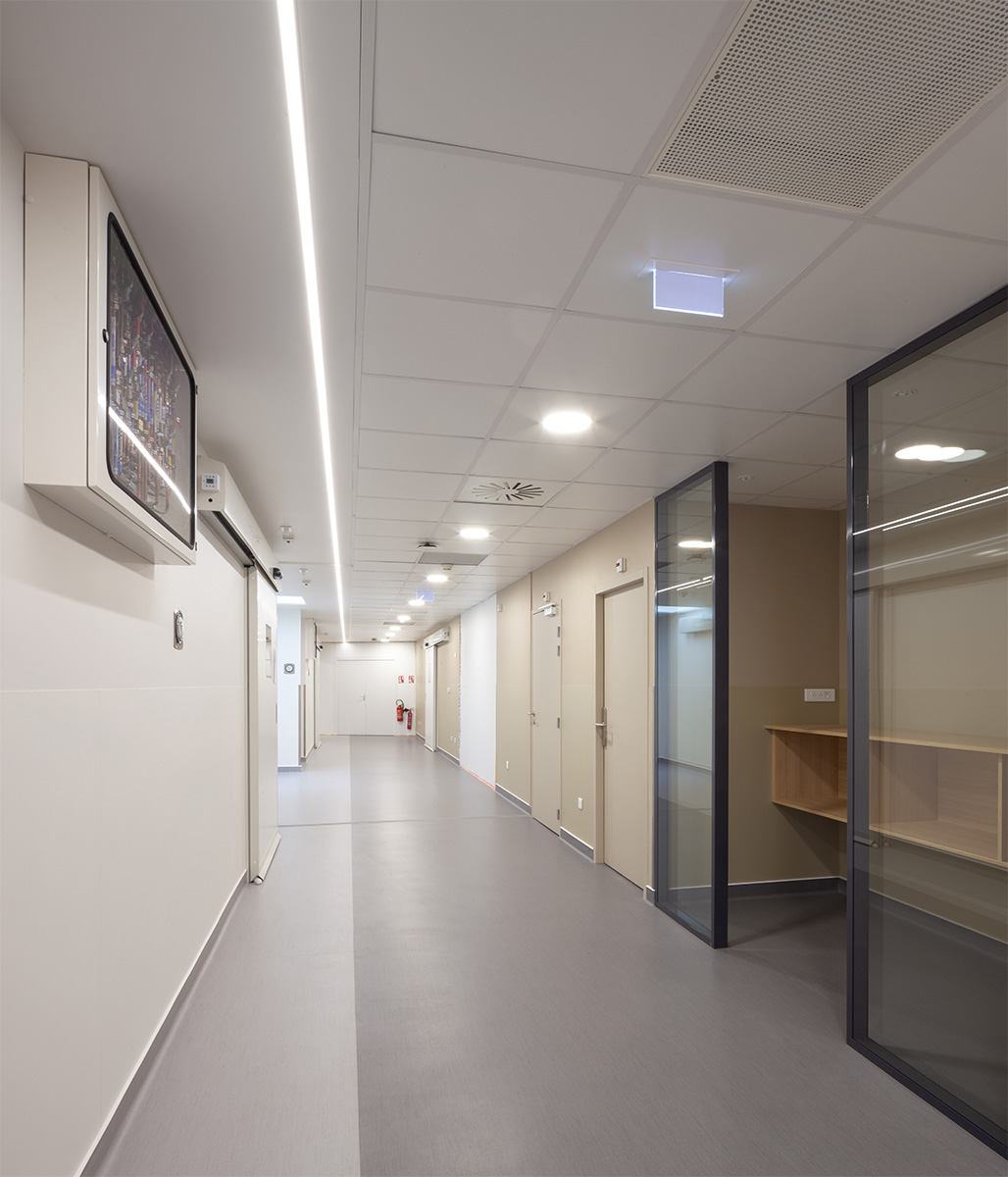 Photographie d'un couloir lumineux blanc et gris, néon lumineux sur toute la longueur et alcôve avec porte vitré coulissante menuiserie gris anthracite