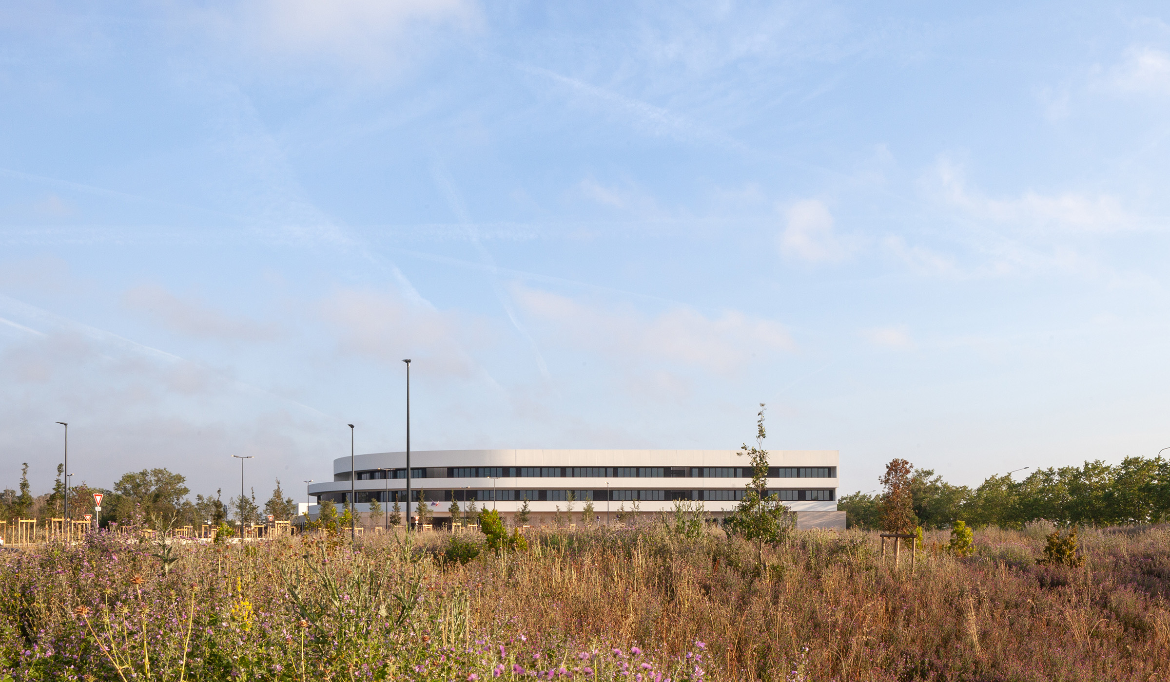 Photographie depuis un champs en friche, au loin on voit la forme du collège avec ses deux fenêtres bandeaux gris sombre, entouré d'un mur auvent blanc