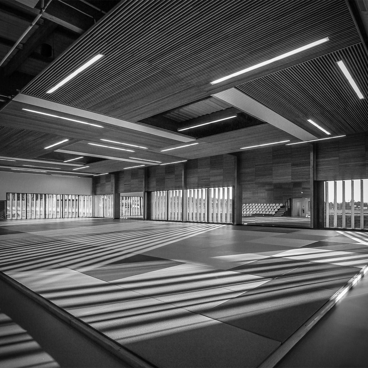 Photographie noir et blanc du dojo vers les baies vitrées et leurs brises soleil, la lumière rasante entre et se reflète sur les miroirs