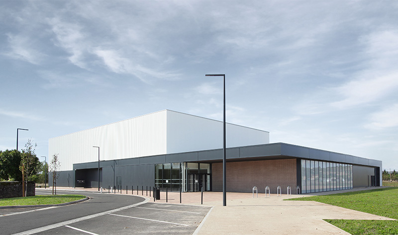 Photographie de l'ensemble du gymnase depuis le trottoir ménant à l'entrée, auvent qui fait le tour du bâtiment gris anthracite et soubassement brique, volume bardage blanc vertical pour le terrain couvert du gymnase