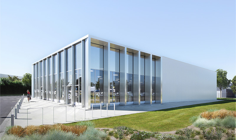 Photographie sur le côte de la façade d'entrée du bâtiment en métal blanc, entrée du bâtiment en vitrage tout hauteur, quelques massifs végétaux au 1er plan