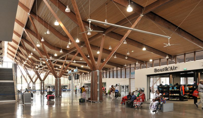 Photographie de l'intérieur du bâtiment vers l'ouverture lumineuse au plafond, des poteaux type quadripode en bois soutiennent la toiture, des gens attendent sur des bancs