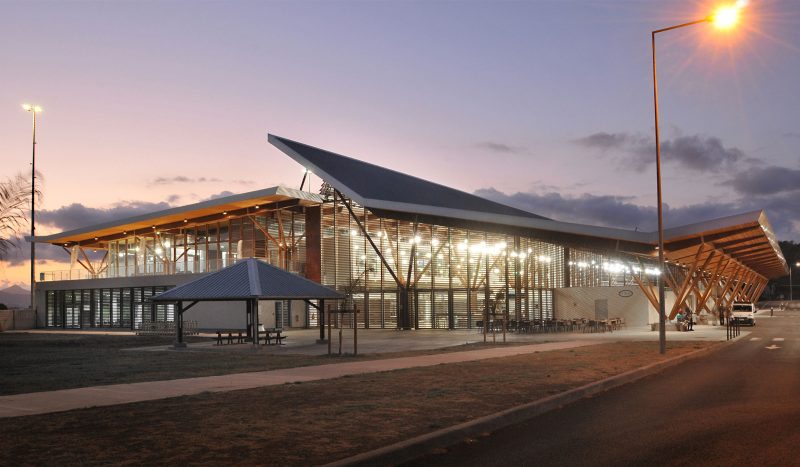 Photographie éloigné de l'aéroport en fin de journée et éclairage de l'intérieur de la structure des poteaux et ventelles en bois, toiture ouverte au milieu et remontante sur le auvent