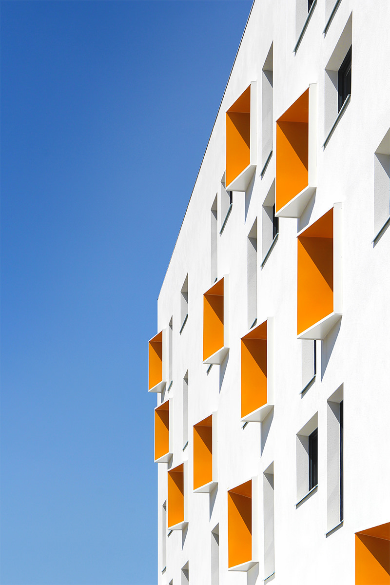 Photographie du soleil éclairant la façade blanche et et ombrant les brises-soleil orange