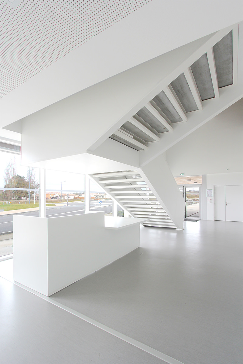 Photographie du hall d'entrée du gymnase tout blanc avec escalier suspendu, constrastant avec le corten