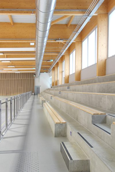 Photographie verticale de l'intérieur du gymnase au niveau des gradins, béton brut et bois éclairé par un long vitrage bandeau