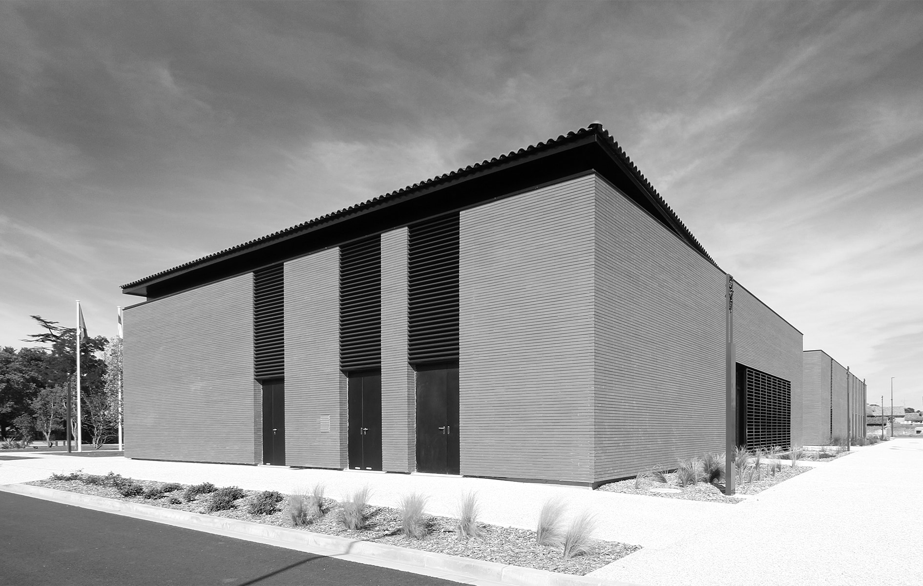 Photographie noir et blanc de trois quart de l'arrière du bâtiment vêtue de briques, volumes rectangulaire avec une ouverture dans la longueur du bâtiment