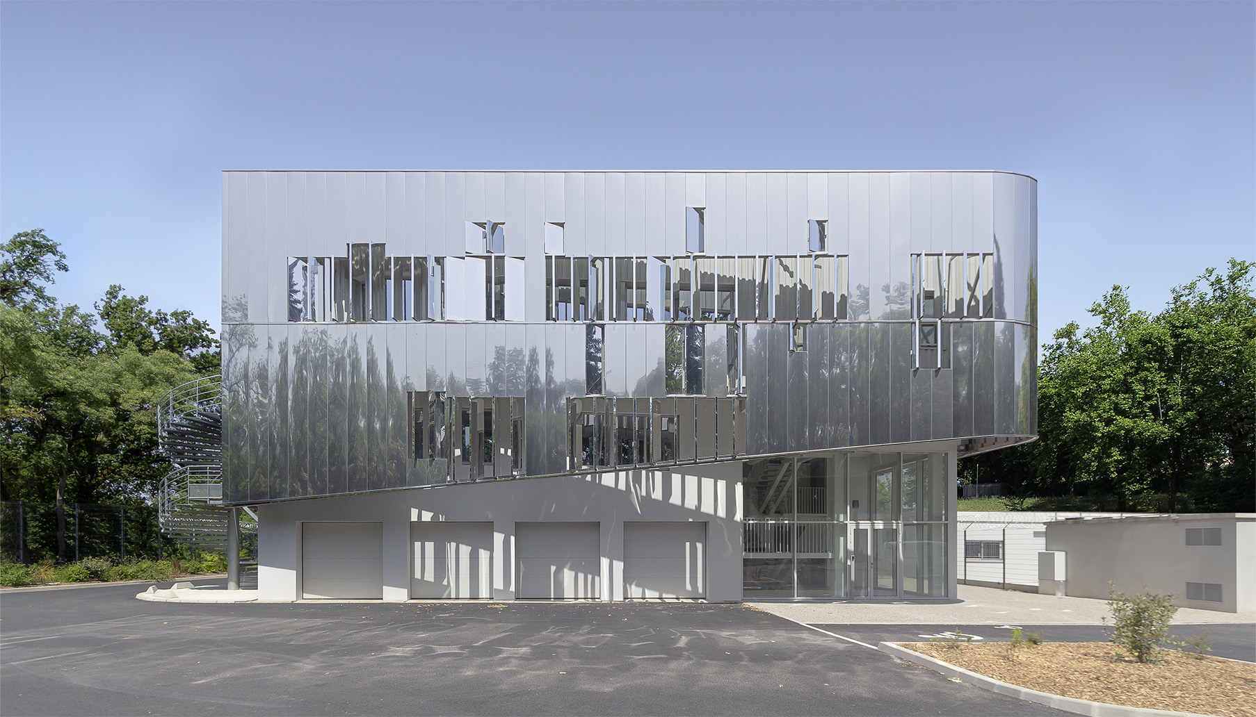 Photographie de la façade reflétant la fôret au alentours, soubassement béton et enveloppe miroir