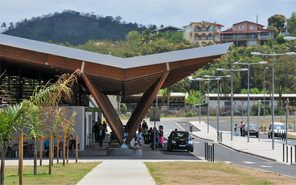 Photographie de l'enfilade de poteaux portant un grand auvent protégeant plusieurs personnes du soleil sur le parvis de l'aéroport de Mayotte