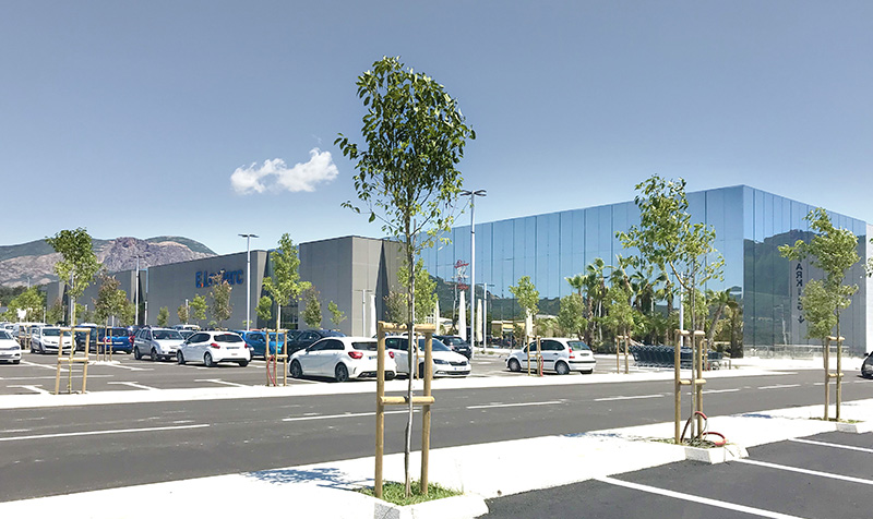 Photographie depuis le parking du centre commercial, façade alternance de métal perforé gris et de miroir tout hauteur réfletant les montagnes