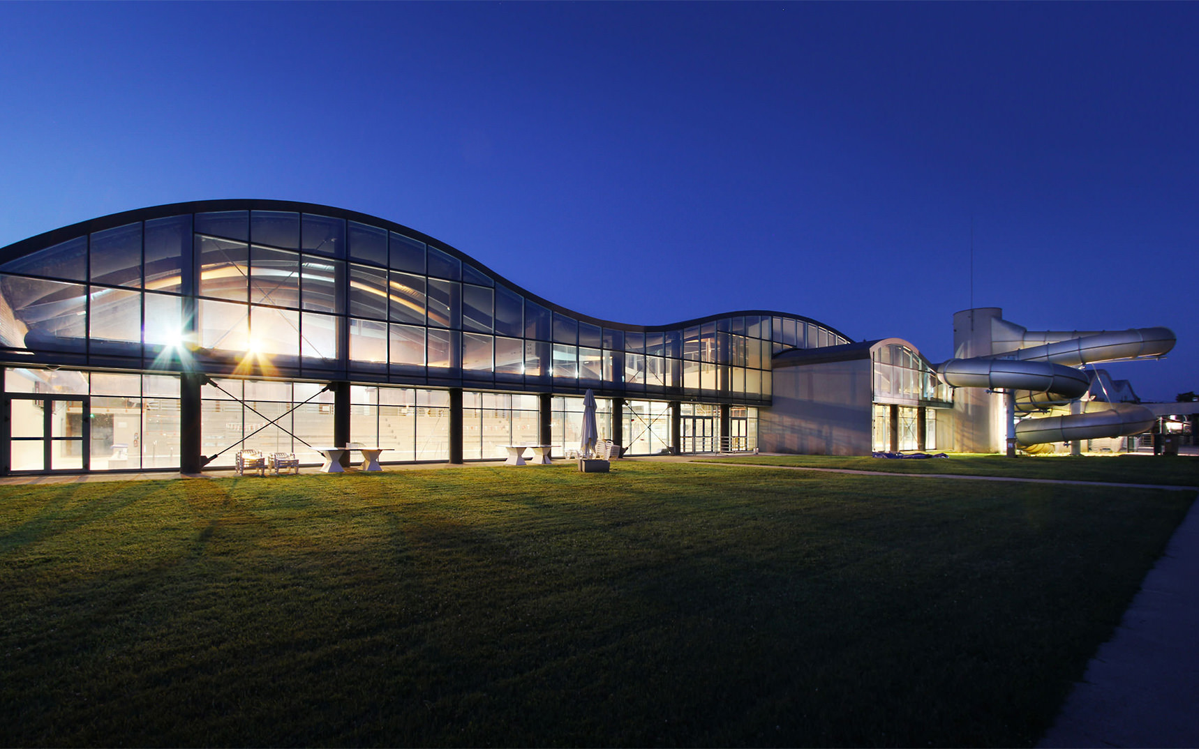 Photographie depuis le cheminement extérieur du complexe aquatique de nuit éclairé de l'intèrieur avec le vitrage tout hauteur