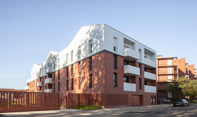 Photographie depuis la rue Jean Jaurès à Montauban, vue sur le côté du bâtiment R+3, brique et moucharabieh en bas, balcons, dernier étage et toiture en blanc, triple toiture pente douce