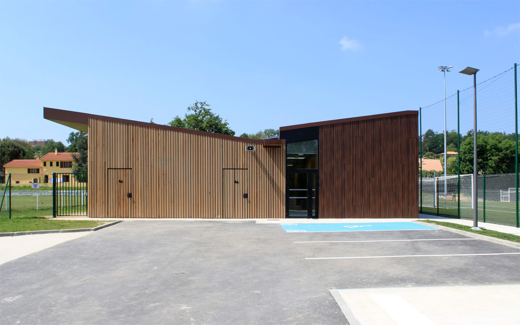 Photographie depuis le parking de la façade opposée à l'entrée du bâtiment, lames en bois et toiture avec pente vers le centre du bâtiment