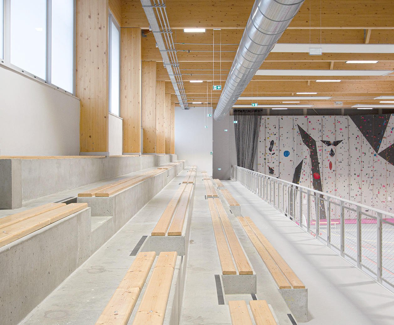 Photographie intérieure depuis les gradins qui surplombent les terrains de sport, gradins béton et bois, au plafond poutres bois traversantes apparentes
