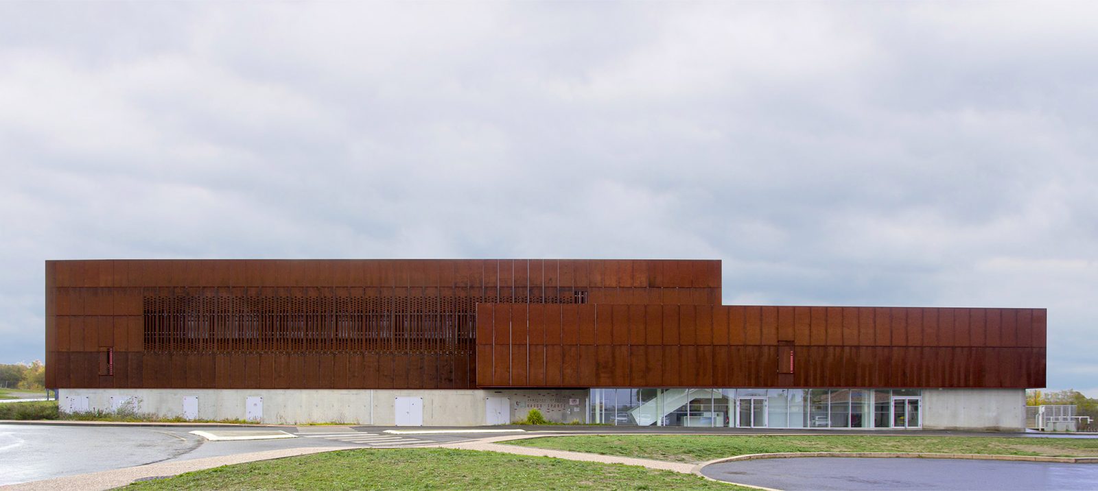  Photographie de la façade principale depuis l'espace vert du parking, deux volumes rectangulaires s'unissent avec un même soubassement béton et un même matériau corten mais n'ont pas la même hauteur