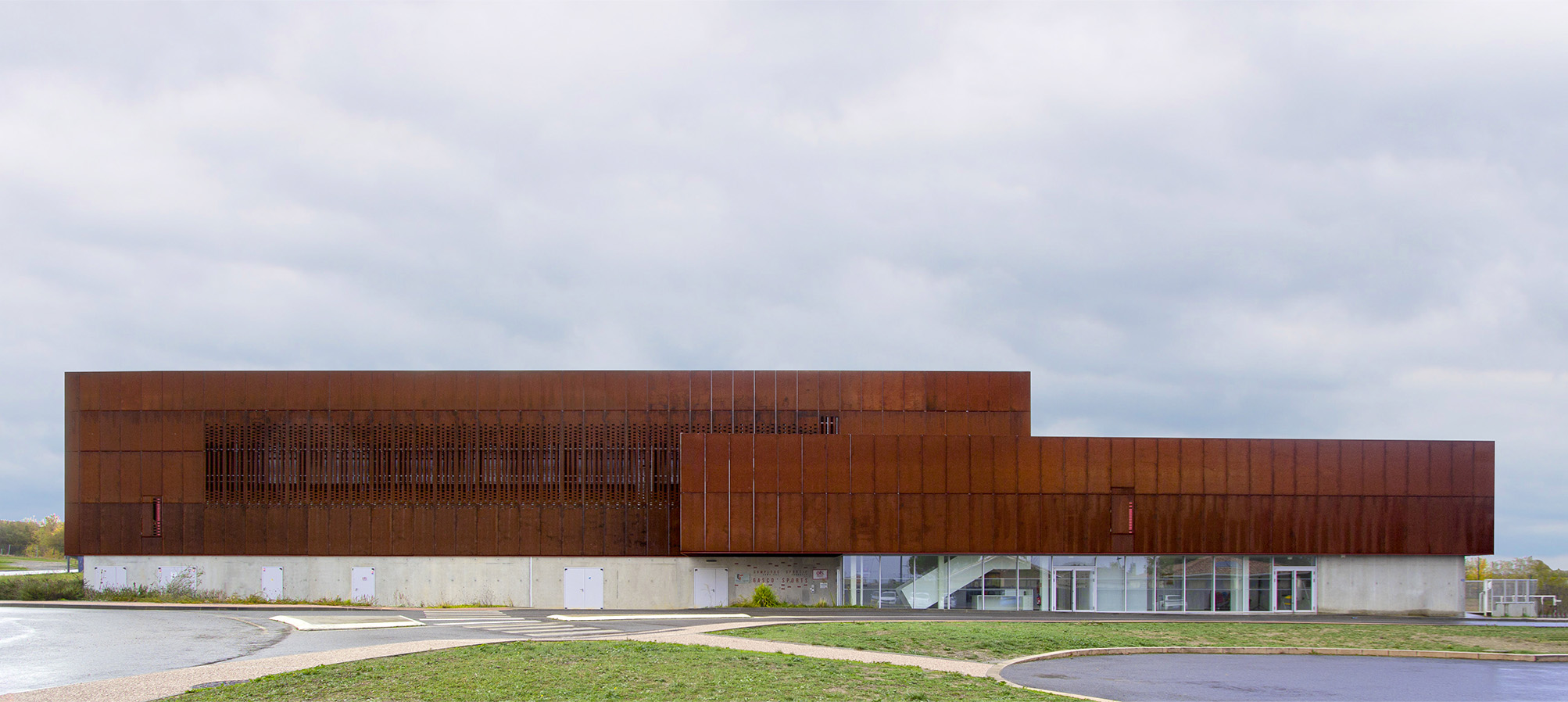 Photographie de la façade principale depuis l'espace vert du parking, deux volumes rectangulaires s'unissent avec un même soubassement béton et un même matériau corten mais n'ont pas la même hauteur