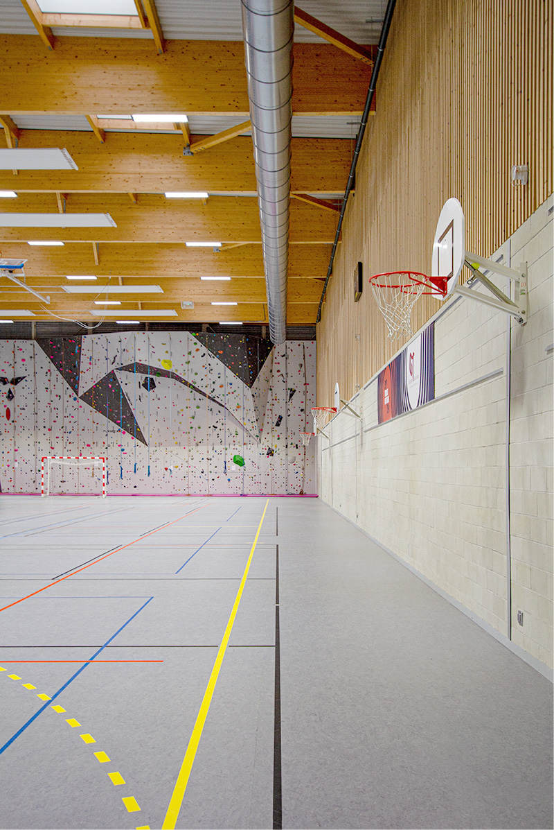 Photographie au niveau des terrains de jeu, face au mur d'escalade tout hauteur, mur à droite, en lame bois vertical et soubassement béton, éclairé par des puits de lumiéres en enfilade