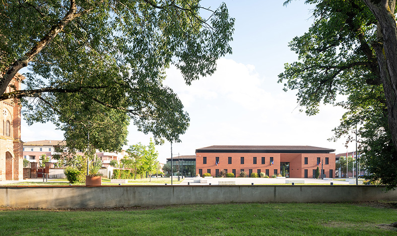 Photographie depuis un espace arboré au loin prés de l’église sur le coté, le bâtiment est entre deux arbres, herbe au 1er plan