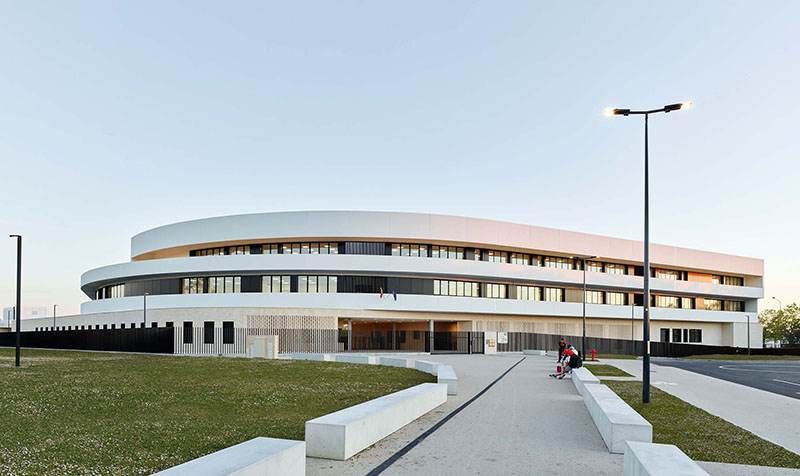 Photographie depuis l'allée centrale vers l'entrée du collège, alterance de bandeaux vitrée gris anthracite cerné d'un mur auvent blanc sur 2 niveaux en escalier