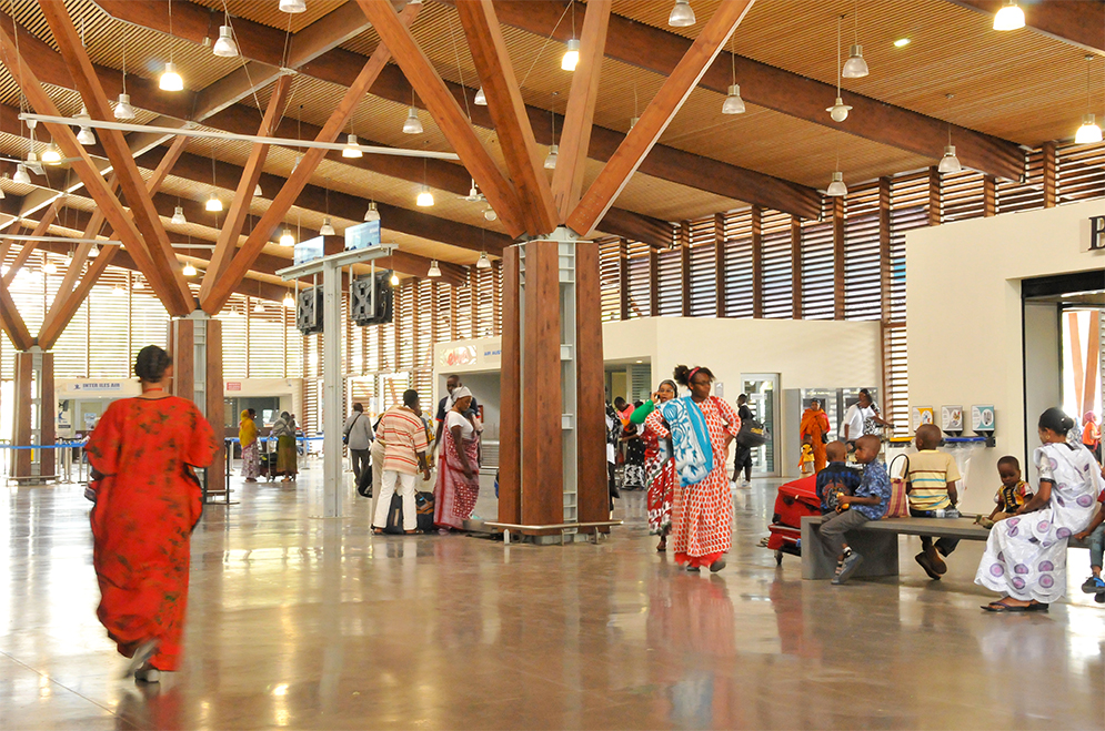 Photographie du hall avec succession de poteaux bois en étoile, plafond et poutres bois, sol en béton lisse refléchissant, plusieurs personnes circulent et attendent sur des bancs
