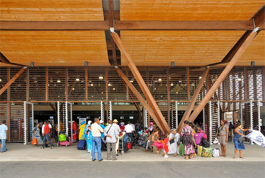 Photographie en face de plusieurs entrées de l'aéroport de Dzaoudi-Pamandzi, façade en lamelles bois, poteaux en étoile et auvent en bois