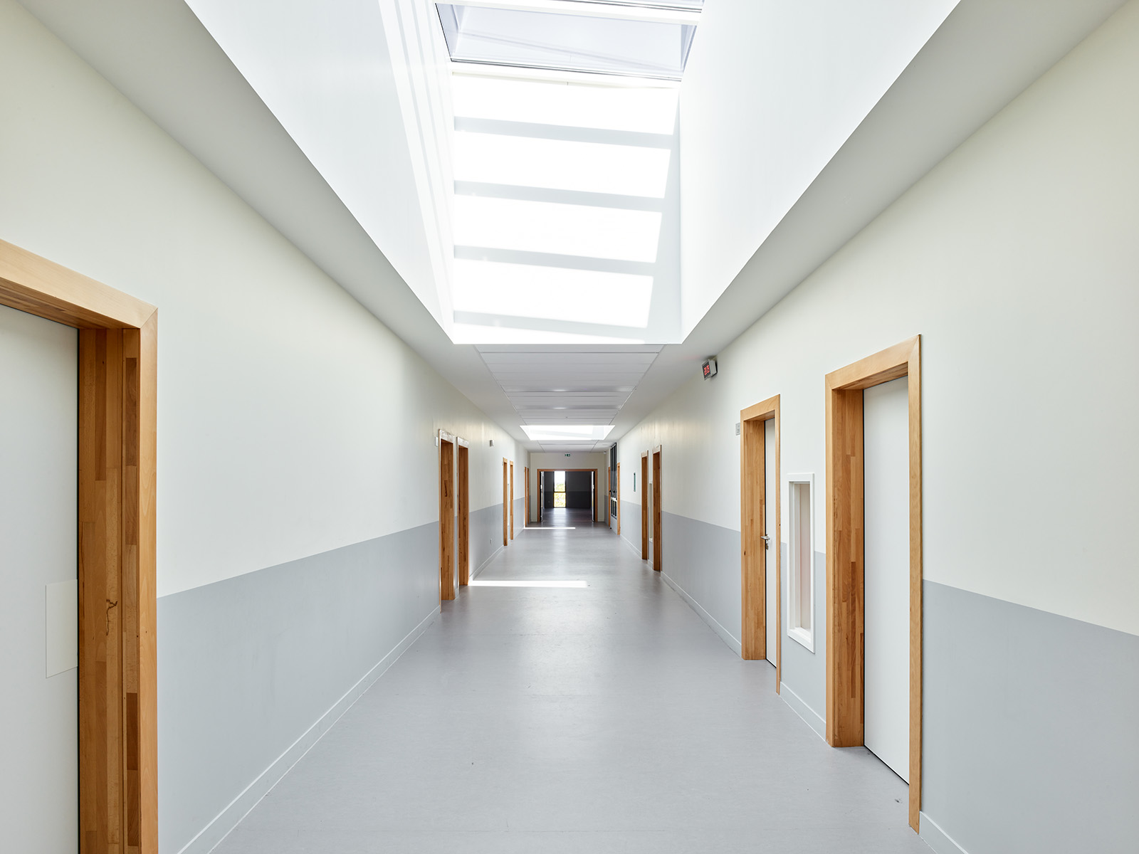 Photographie face à un couloir blanc et partie basse avec le sol gris clair, enfilade de portes menuiserie bois, en hauteur des puits de lumière apporte de la luminosité