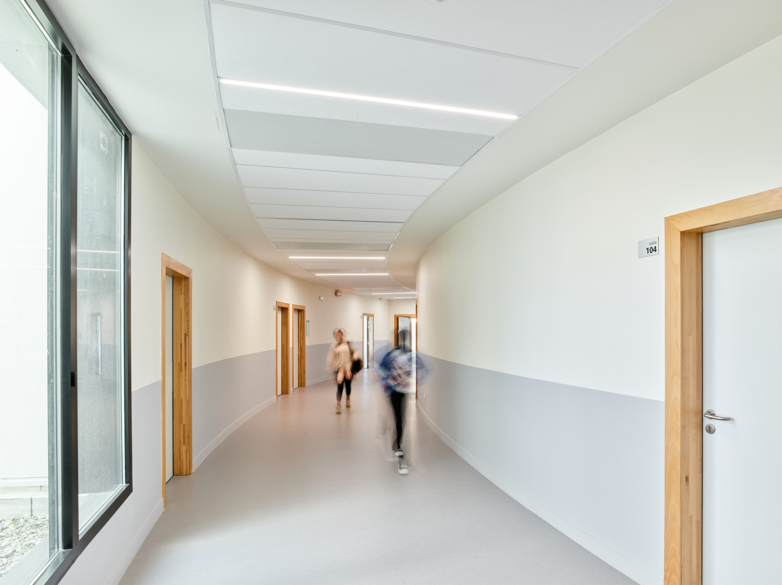 Photographie face à la courbe d'un couloir blanc et gris clair en partie basse et menuiserie bois, des collégien·nes marchent dans un mouvement flou
