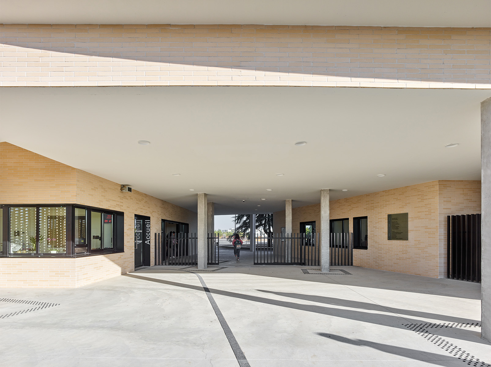 Photographie face aux grilles d'entrées à côté du local d'accueil, plafond blanc et soubassement brique claire , le passage traverse le bâtiment et on peut voir en fond la cour avec une partie d'un cyprès