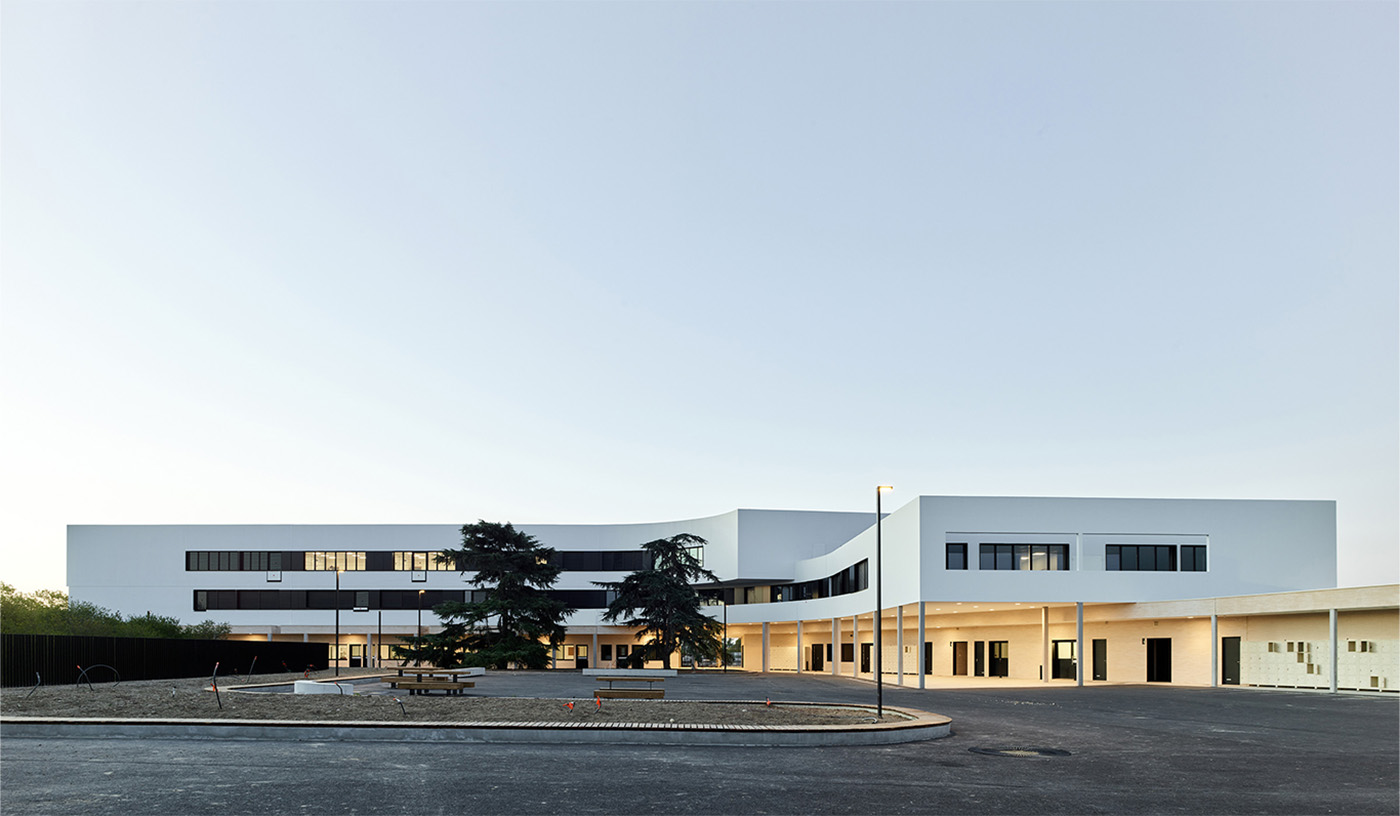  Photographie depuis la cour du collège du bâtiment blanc et sa courbe entourant les deux cèdres en face, fenêtres bandeaux sombres sont éclairés dans certaines salles qui commencent à prendre vie, éclairage jaune sous le préau et ciel trés clair